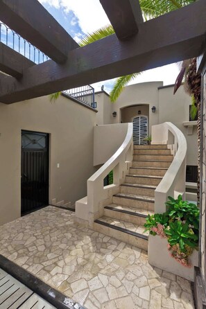 Eine Treppe führt zu einer Tür mit einem Metallgitter, einer mit Steinplatten belegten Terrasse und einem Pergolen-Aufbau.