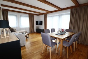 Ein modernes Wohnzimmer mit Holzbalkendecke, einem Flachbildfernseher, einem Esstisch mit Stühlen, einer Couch und einem Couchtisch.