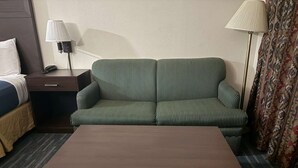 Quarto de hotel com sofá verde listrado, mesa de centro de madeira, tapete estampado, mesa de cabeceira com abajur e cortina padronada.