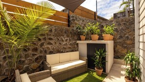 Een terras met een stenen muur, een rieten bank, een kleine vuurkuil en potplanten.