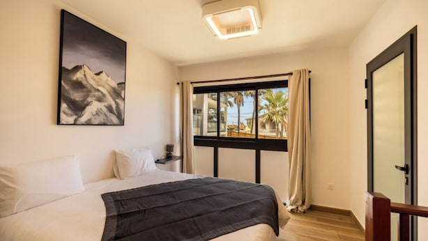 Hotelkamer met een bed, een berglandschap op een schilderij, een raam met uitzicht op palmbomen en een deur met ruit.