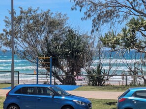 Une zone côtière où une voiture bleue est garée près d’une plage, d’une aire de jeux, et d’où l’on a une vue sur l’océan.