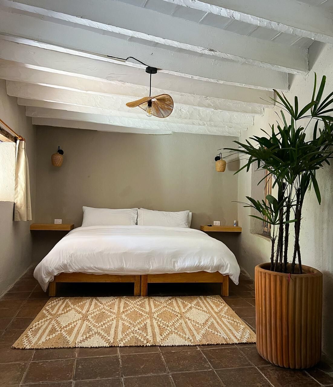 A bedroom with a bed, bedside tables, a hanging light fixture, a large potted plant, and a textured rug.