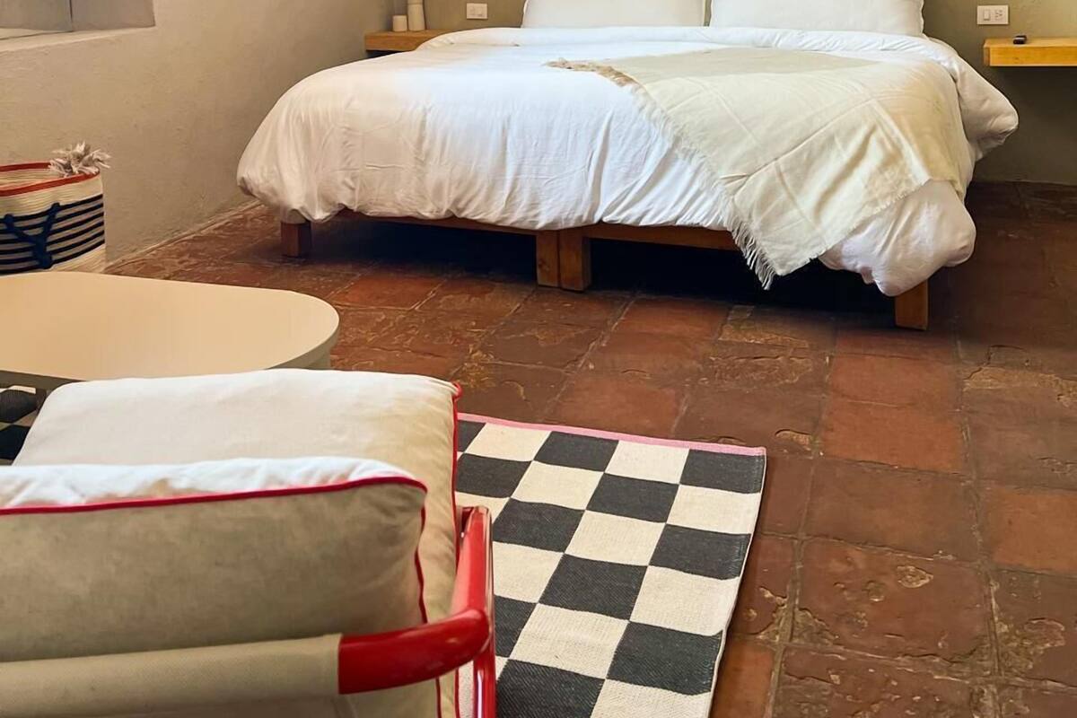 A bedroom with a bed, a small table, a checkered rug, and a window.