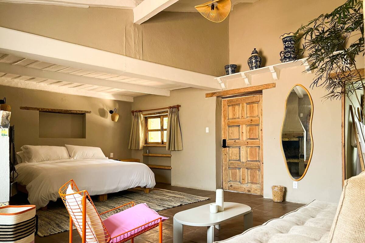 A bedroom with a bed, a small table, a chair, a sofa, a window with curtains, a mirror, and a shelf with decorative items.