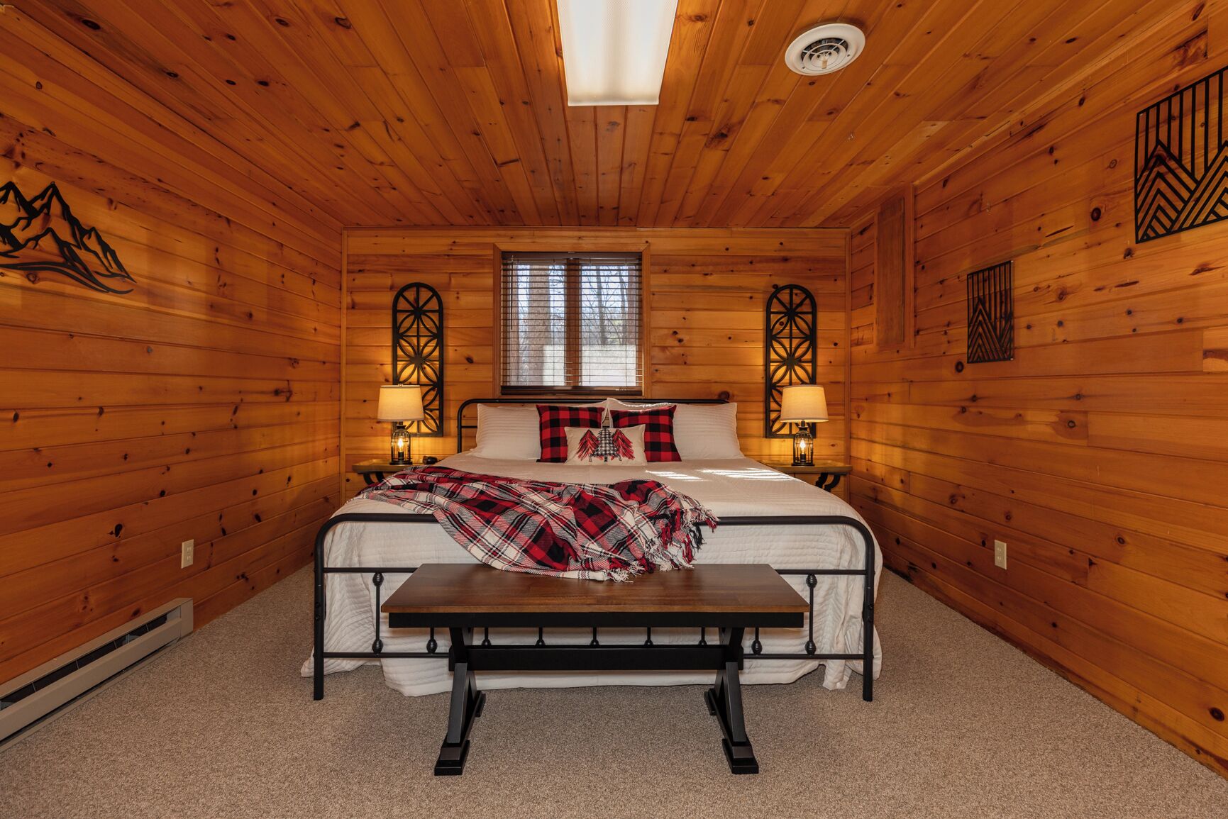 Un dormitorio rústico de madera con una cama, dos mesitas de noche con lámparas, un banco y una ventana con persianas.