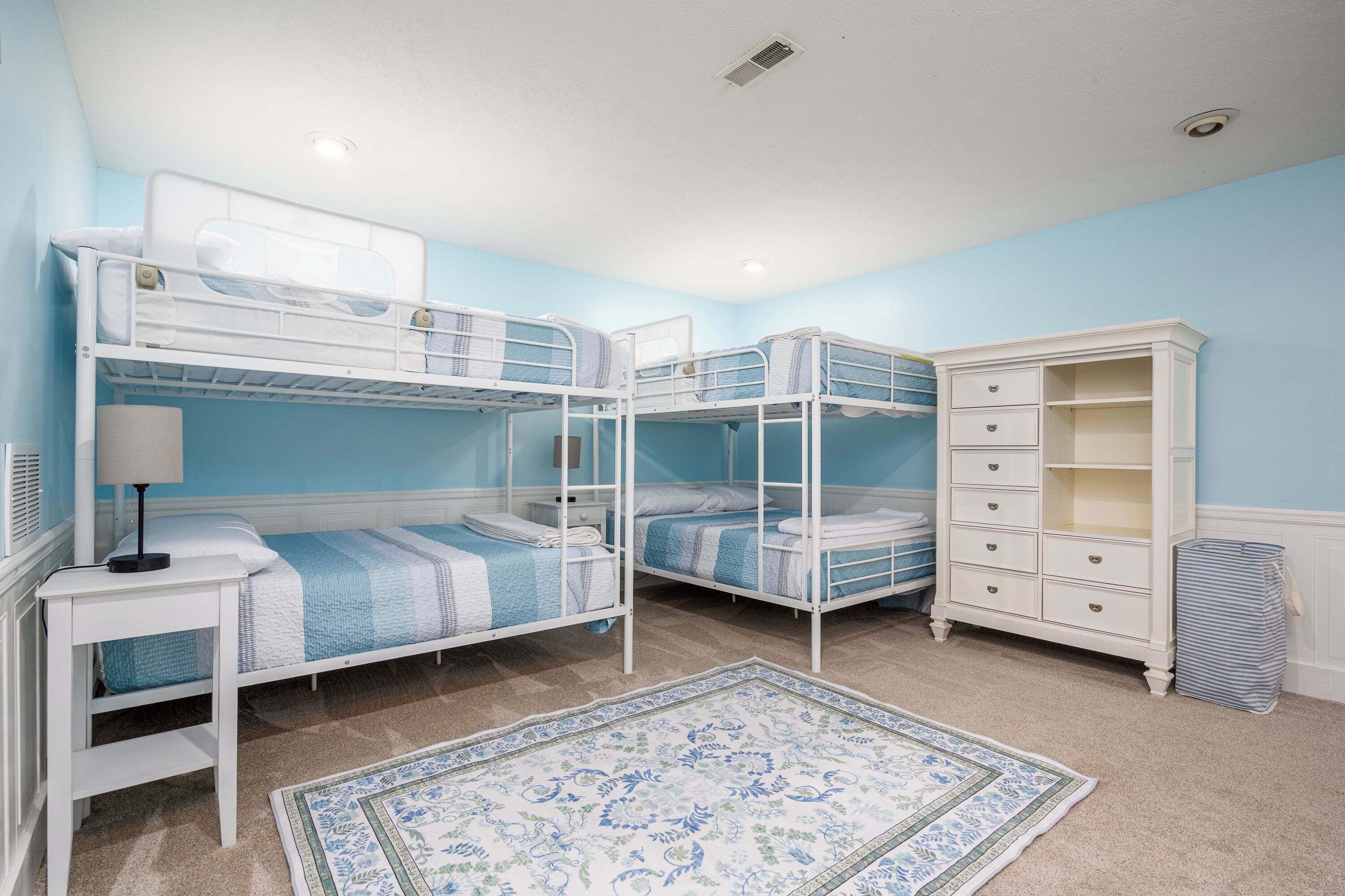 A room with bunk beds, a white dresser, a lamp, and a patterned rug.