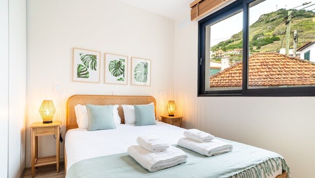 Cama bem arrumada com duas almofadas, uma luminária de cabeceira e uma janela com vista para uma colina.