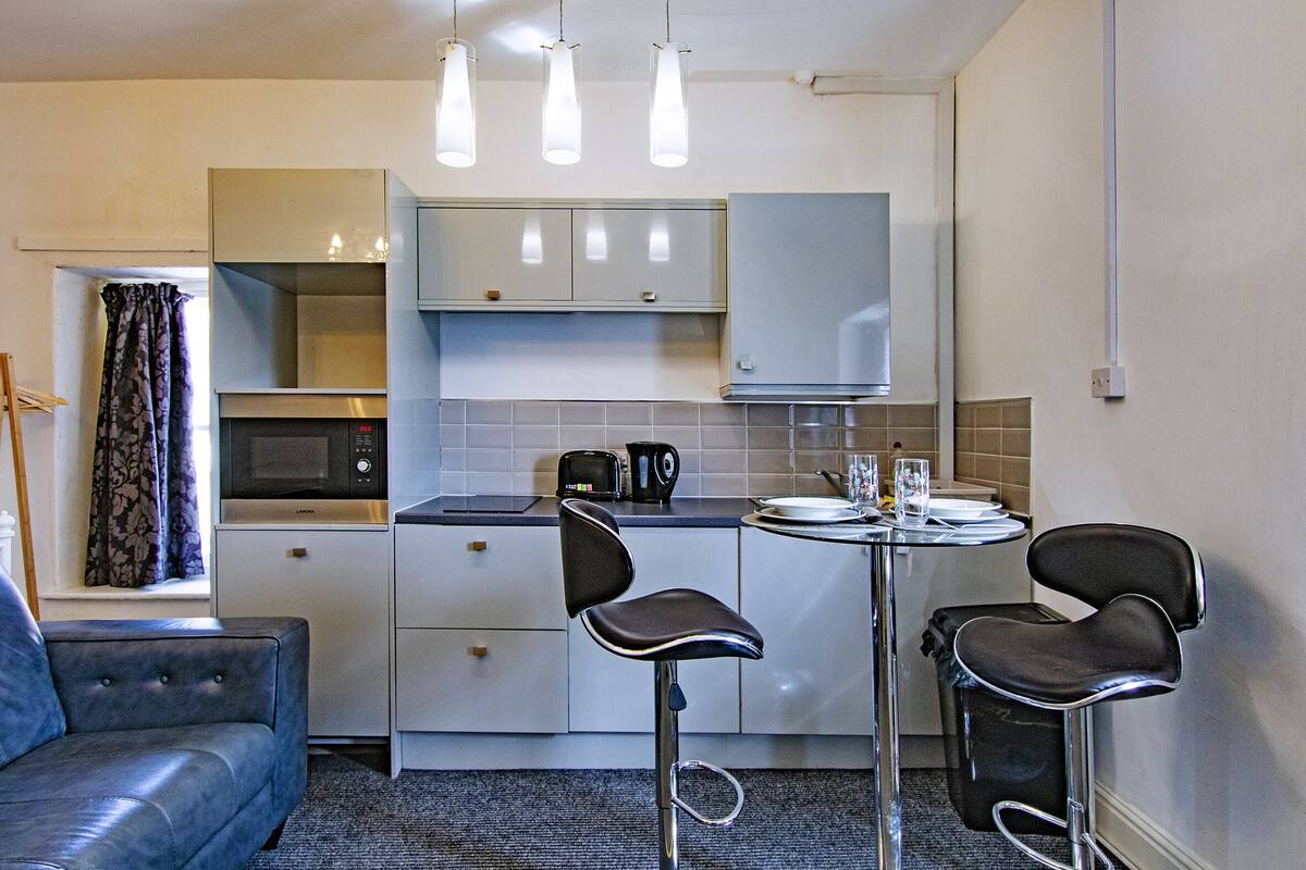 A compact kitchen with a microwave, toaster, and kettle, a small dining table with two chairs, and a sofa.