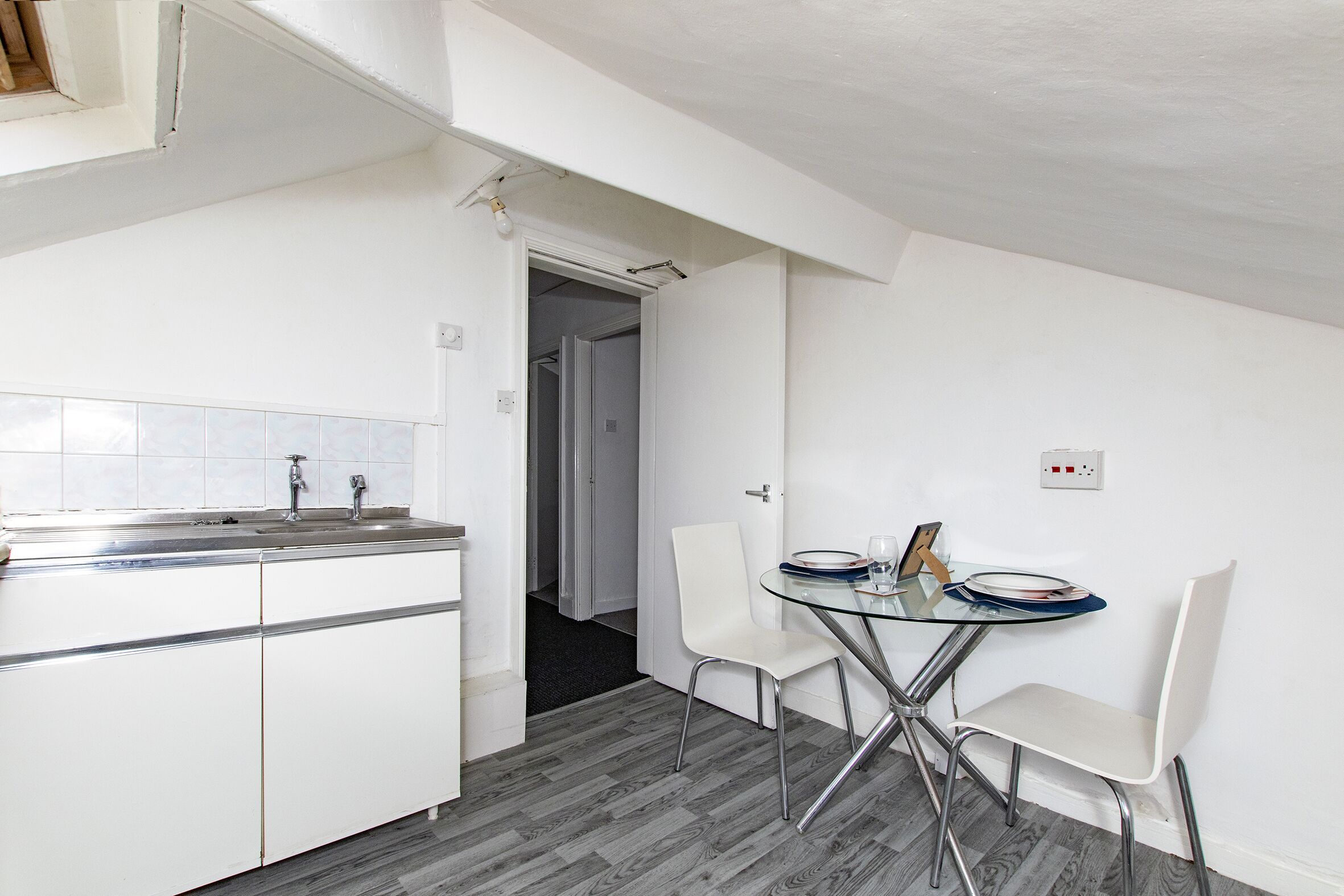 Una cocina compacta con armarios blancos, un lavamanos de acero inoxidable y una mesa de vidrio con dos sillas blancas.