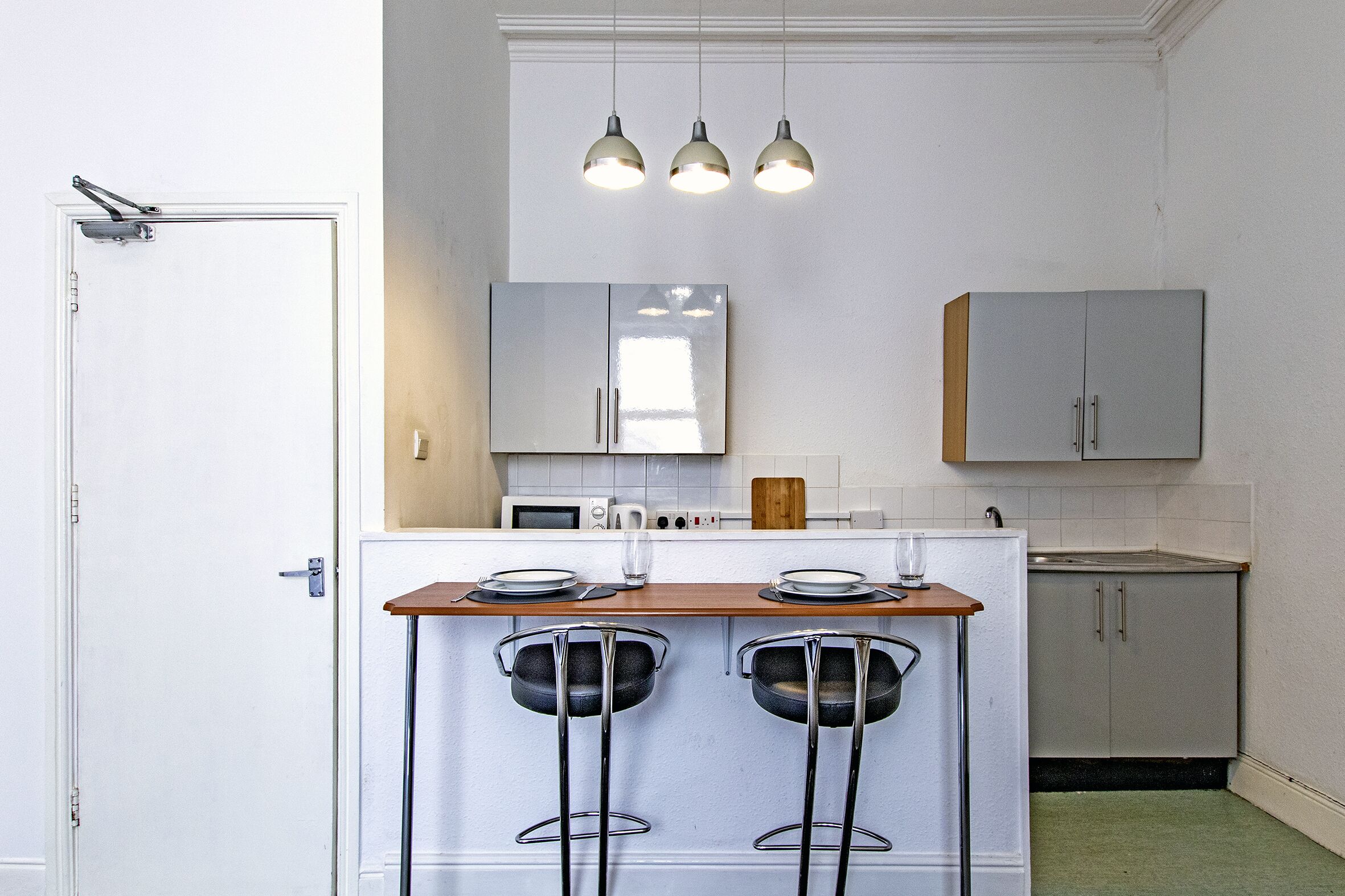 A compact kitchen with a white door, a microwave, a sink, and a double bar stool with a metal frame.