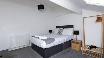 Um quarto com cama, mesas de cabeceira, radiador, luminária pendente e sapateira.
