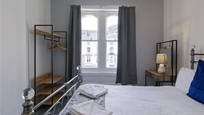 Ett hotellrum med en säng, en hylla i trä, en klädstång, ett nattduksbord med en lampa och utsikt över byggnader genom fönstret.