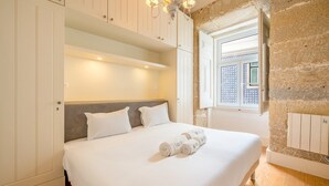 Un dormitorio con una cama grande, ropa de cama blanca, una pared de piedra y una ventana con contraventanas.
