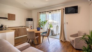 Una cocina moderna con armarios de madera, una zona de comedor con una mesa redonda y sillas, un televisor montado en la pared y un ventanal corredero que da a un balcón.