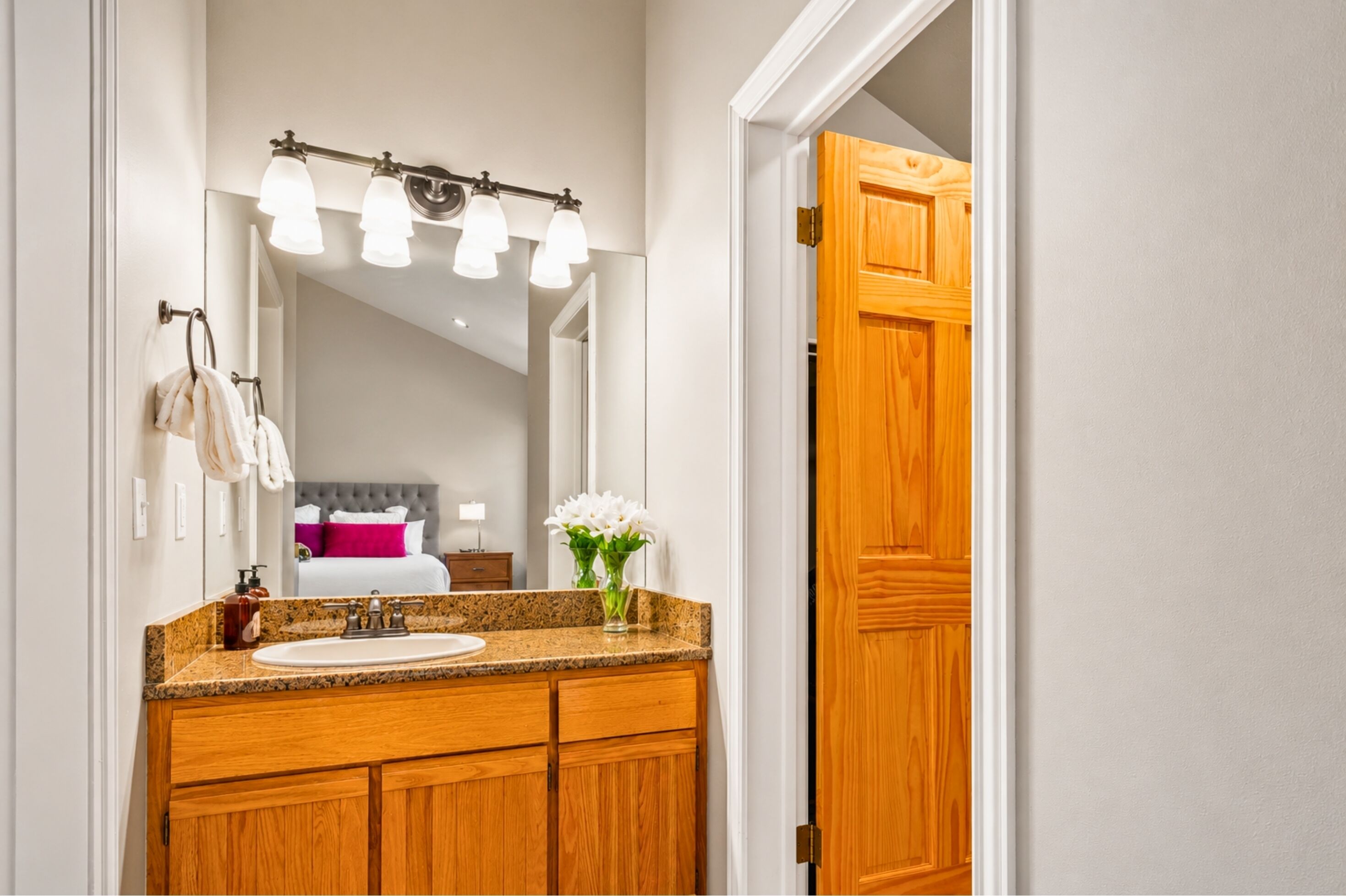 A bathroom with a vanity, sink, mirror, and a bed visible in the background.