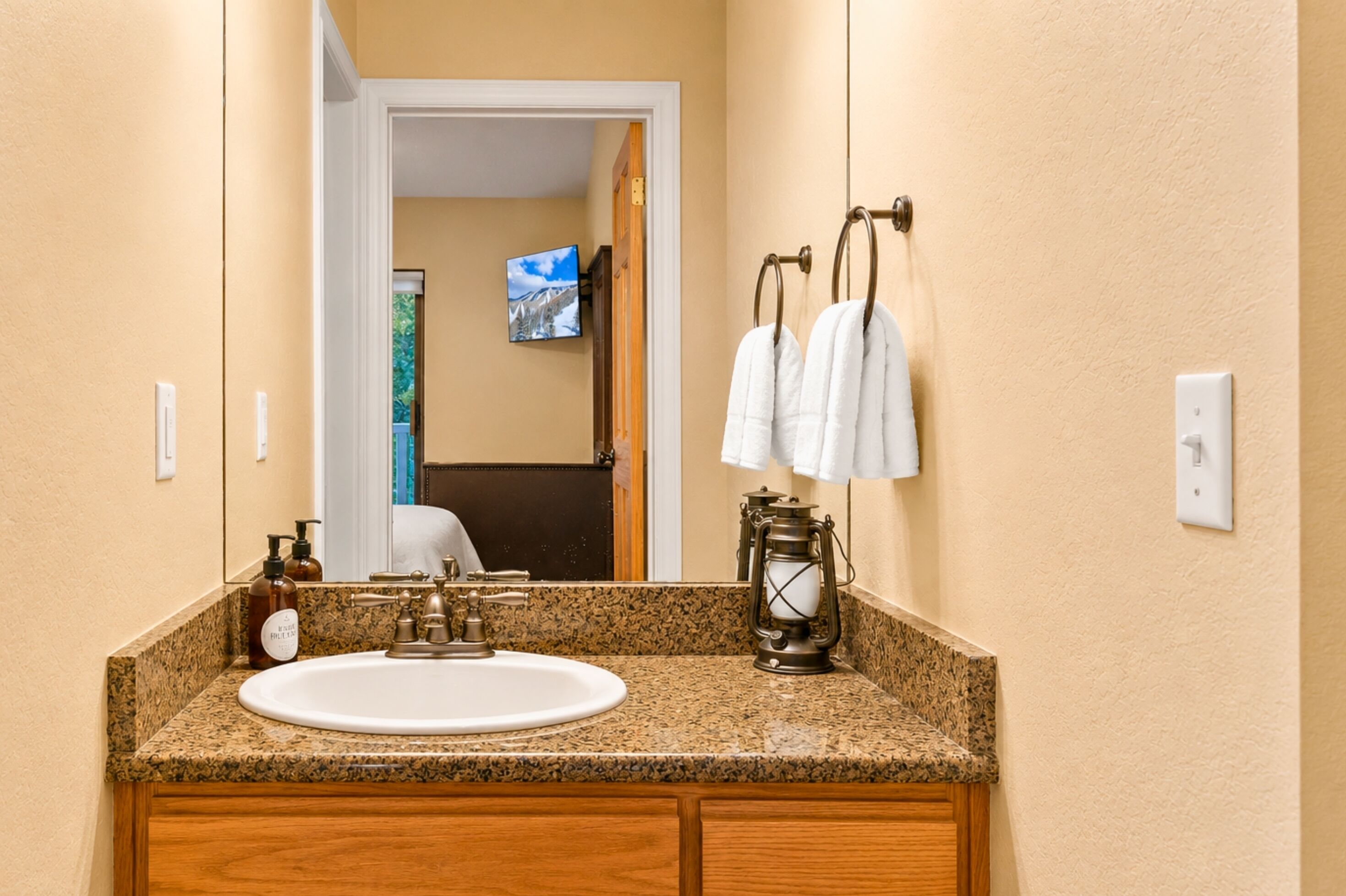 A bathroom with a granite countertop, a white sink, a wooden cabinet, a mirror, a television, and a hanging towel rack.