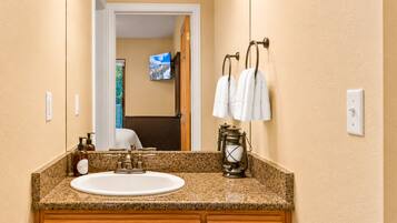 A bathroom with a granite countertop, a white sink, a wooden cabinet, a mirror, a television, and a hanging towel rack.