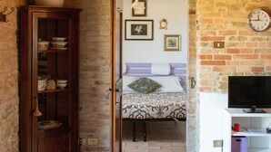 Une pièce au style rustique, avec des murs en briques apparentes, une armoire en bois, un lit recouvert d’une courtepointe à motifs, un téléviseur posé sur un meuble blanc et une horloge au mur.