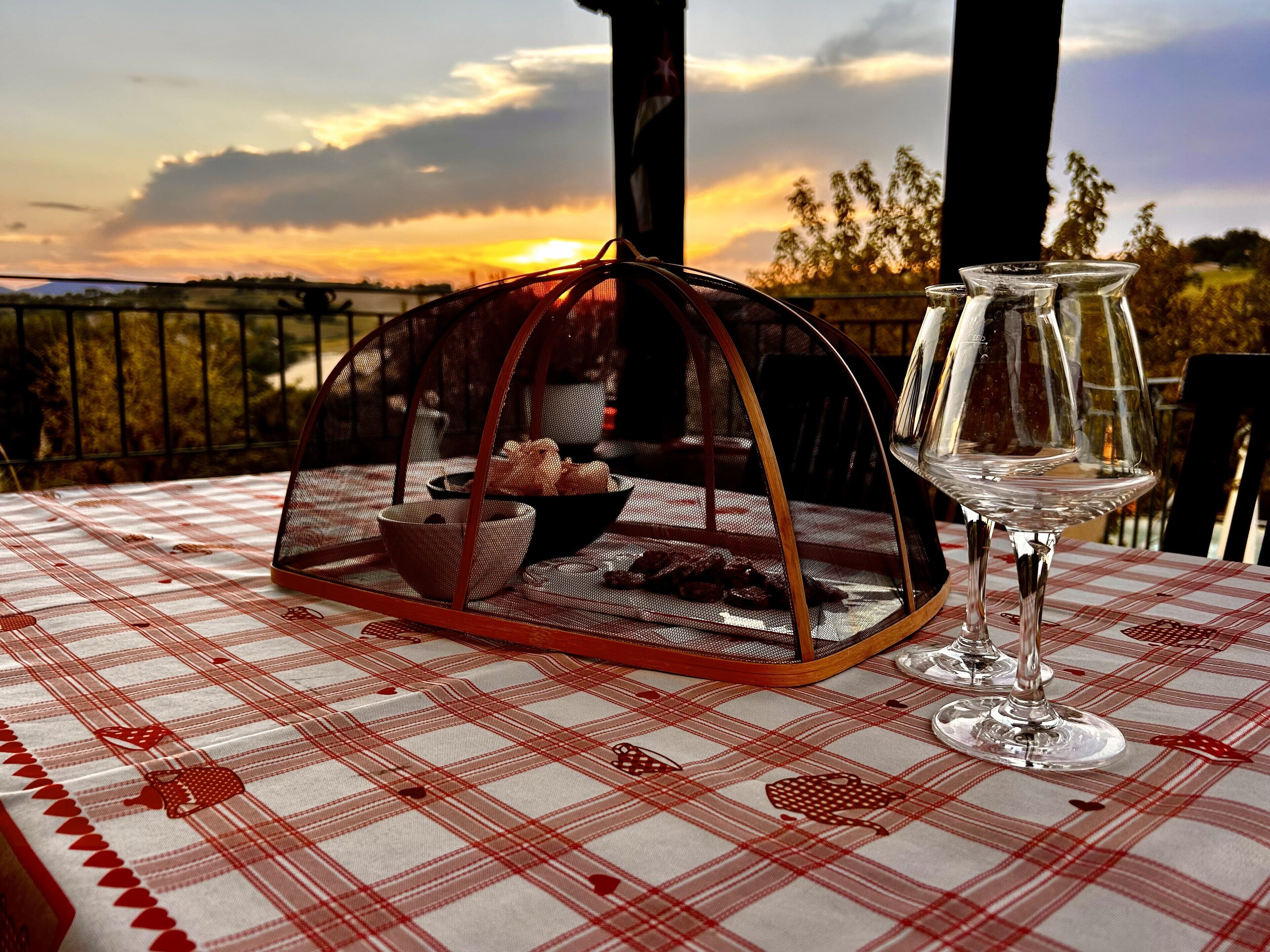A table with a checkered tablecloth, a glass of wine, and a food basket on a balcony with a scenic sunset view.