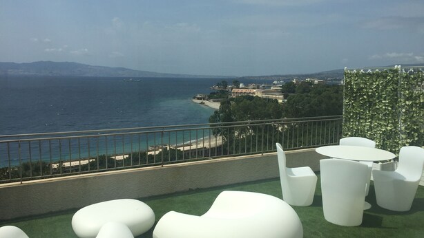 A rooftop terrace with white modern furniture, a view of the sea, and a cityscape in the background. - Reggio Suite Luxury B&B (Reggio Calabria)