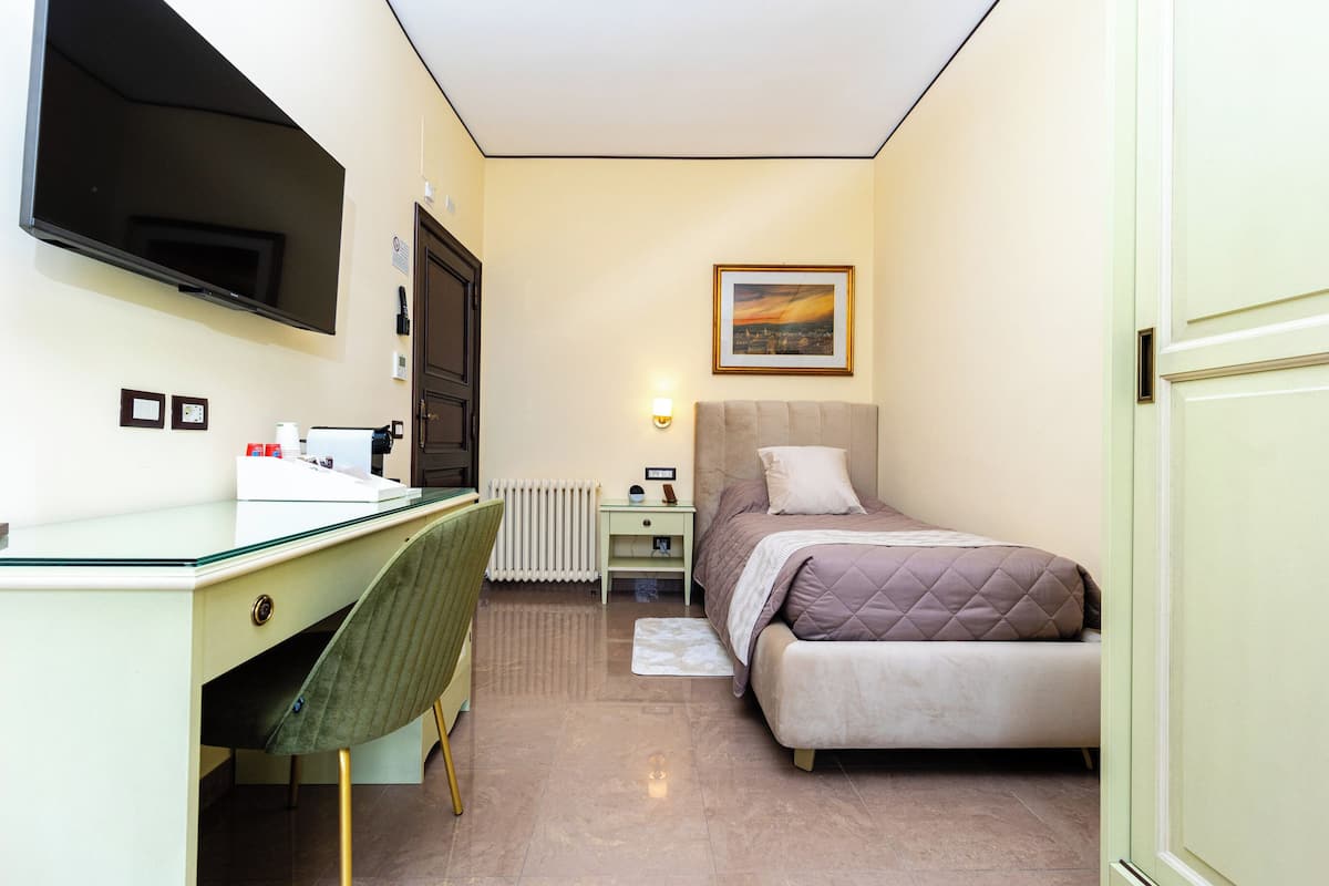 A compact bedroom with a bed, a desk, a chair, a television, and a painting on the wall.