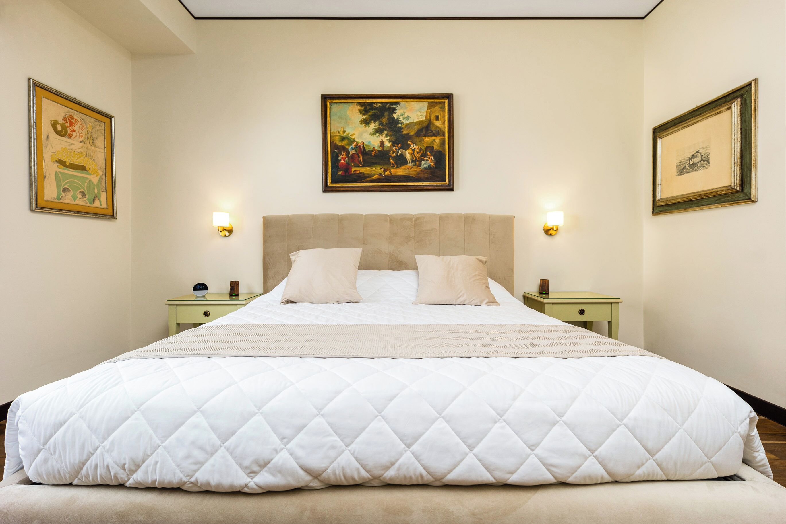 Uma cama grande com edredom branco acolchoado, duas mesas de cabeceira com luminárias e quadros nas paredes.