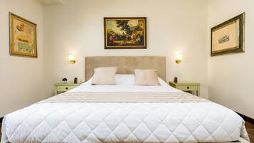 Uma cama grande com edredom branco acolchoado, duas mesas de cabeceira com luminárias e quadros nas paredes.