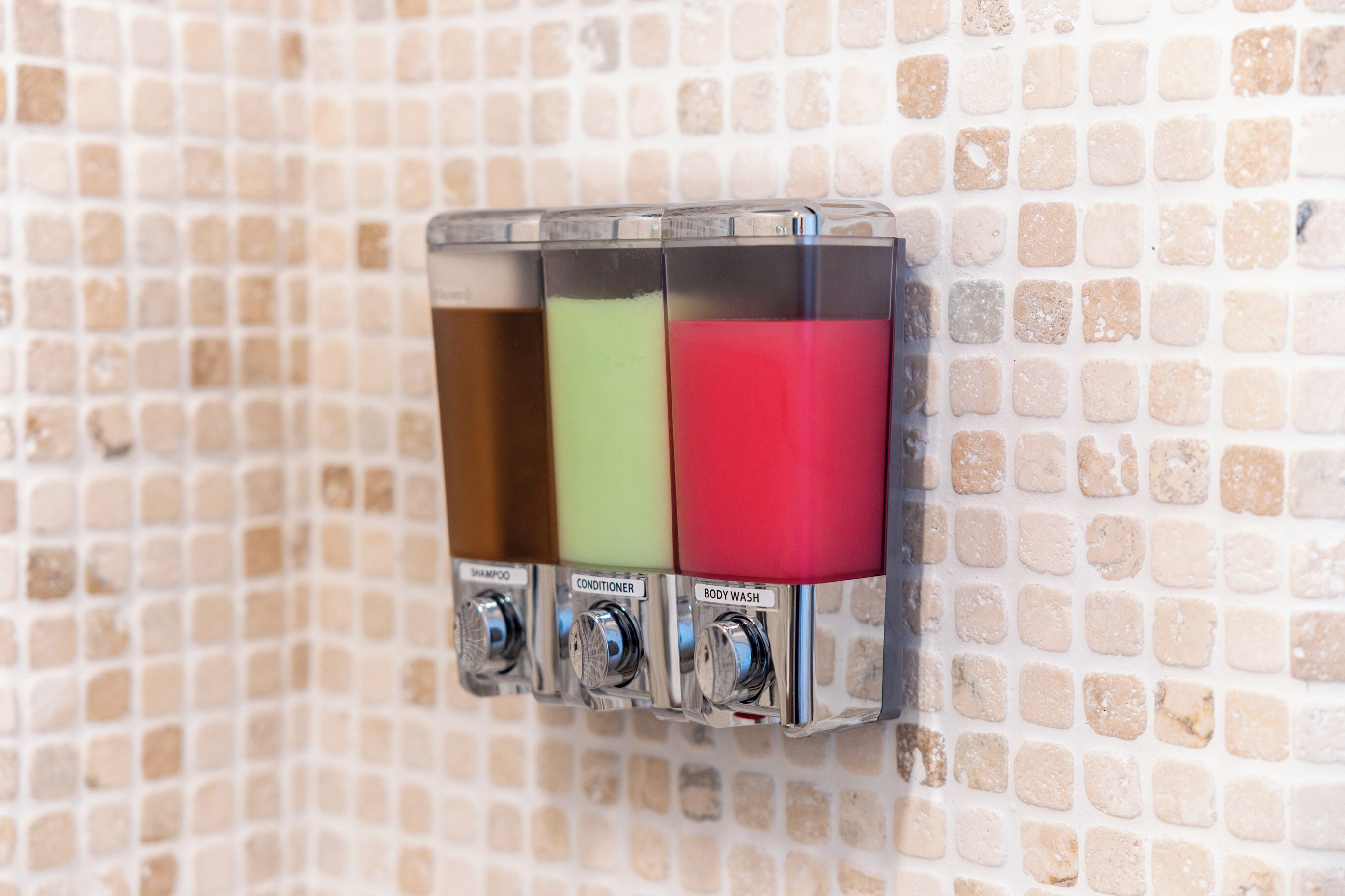 A wall-mounted dispenser with three compartments labeled for shampoo, conditioner, and body wash.