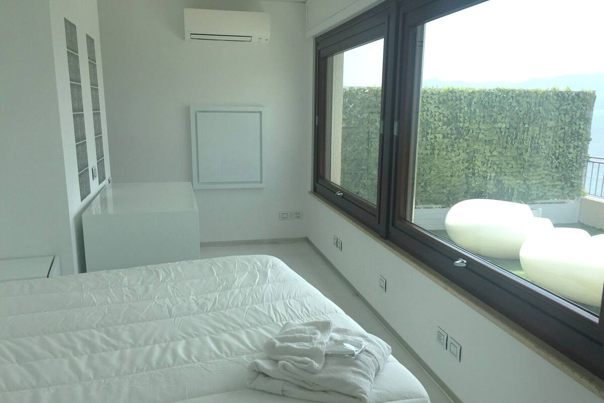 A bedroom with a large bed, a window with a view of greenery, and a white air conditioning unit.