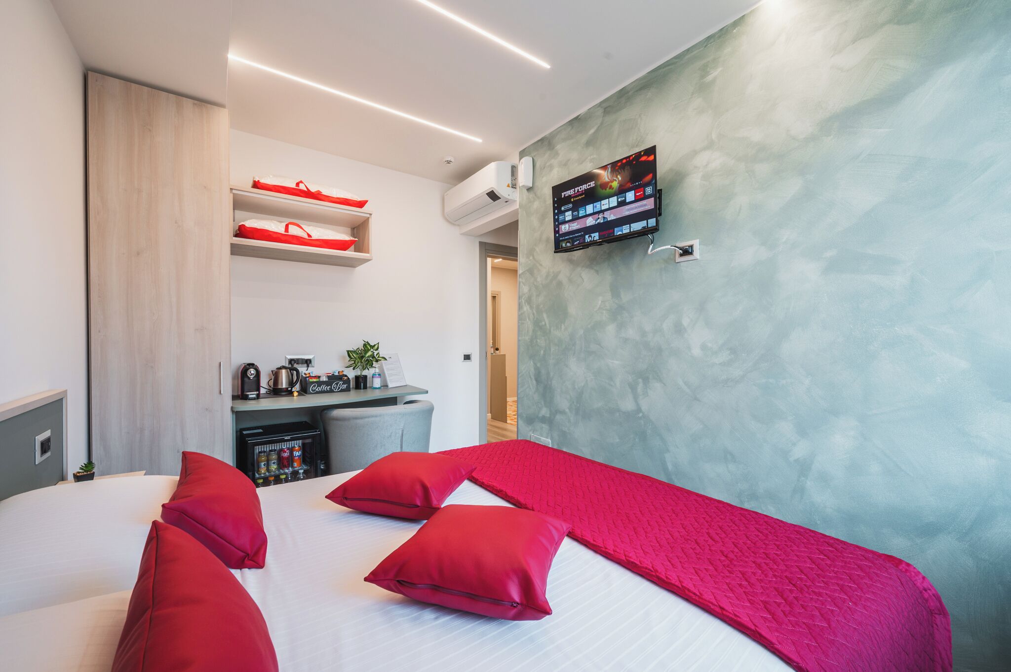 A modern hotel room with a large bed, red pillows, a flat-screen TV mounted on the wall, a minibar, and a small kitchenette area.