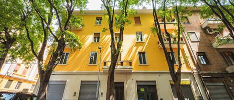 Un edificio giallo con numerose finestre e balconi, auto parcheggiate e alberi di fronte.