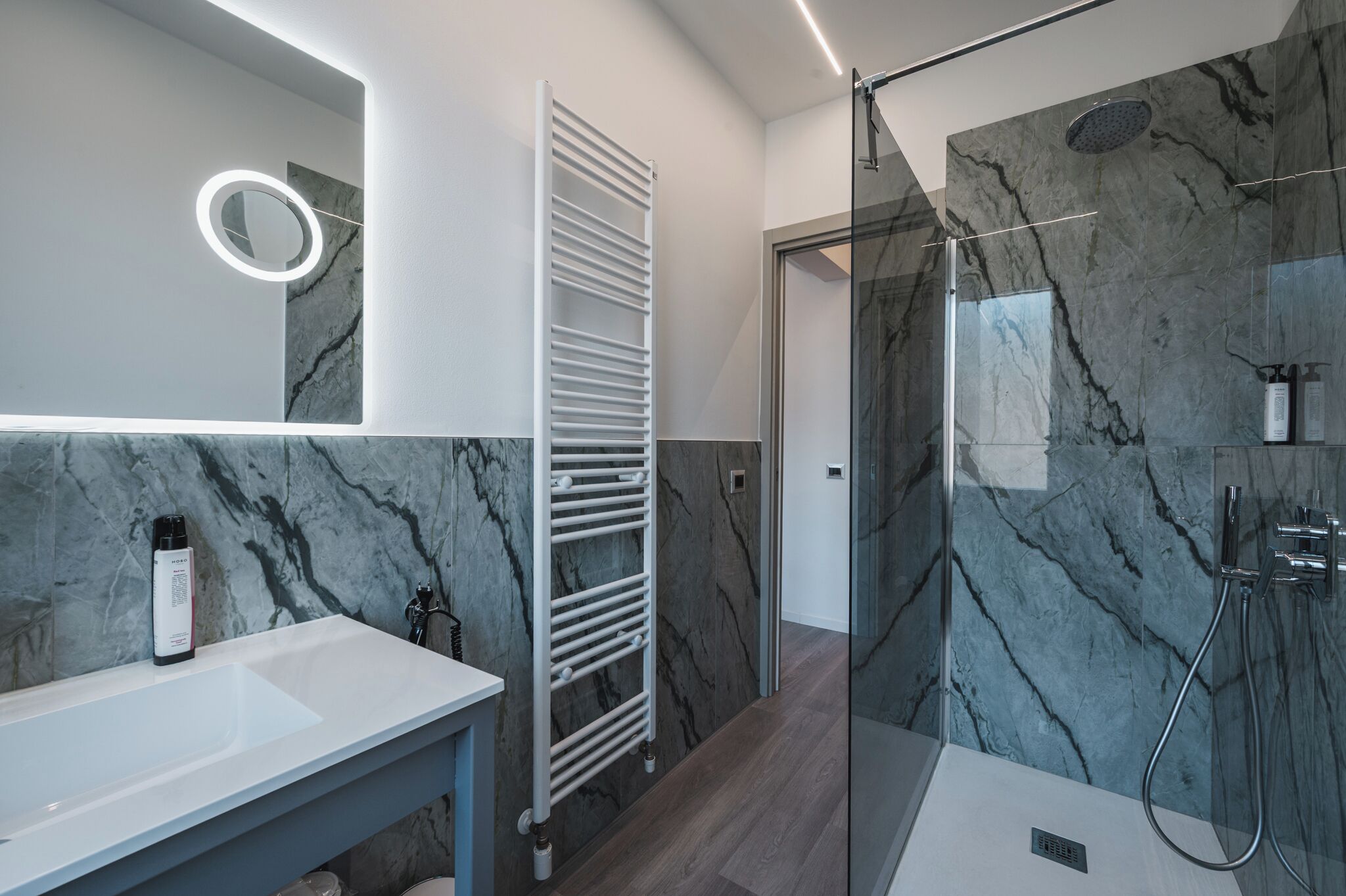 A modern bathroom with a marble backsplash, a glass-enclosed shower, and a white vanity with a mirror and a bottle of shampoo.