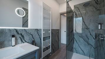 A modern bathroom with a marble backsplash, a glass-enclosed shower, and a white vanity with a mirror and a bottle of shampoo.