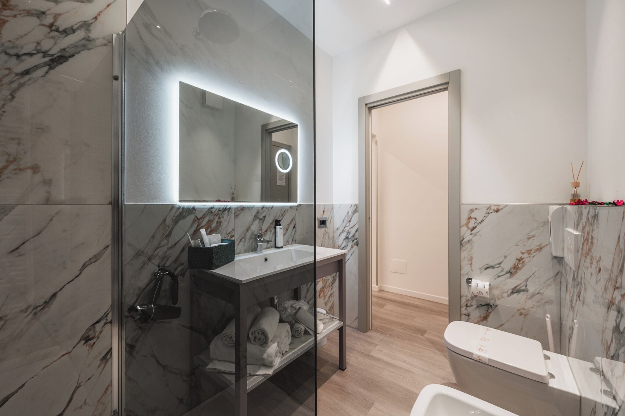Uma casa de banho moderna com chuveiro de vidro, parede revestida a mármore, lavatório com espelho e sanita.