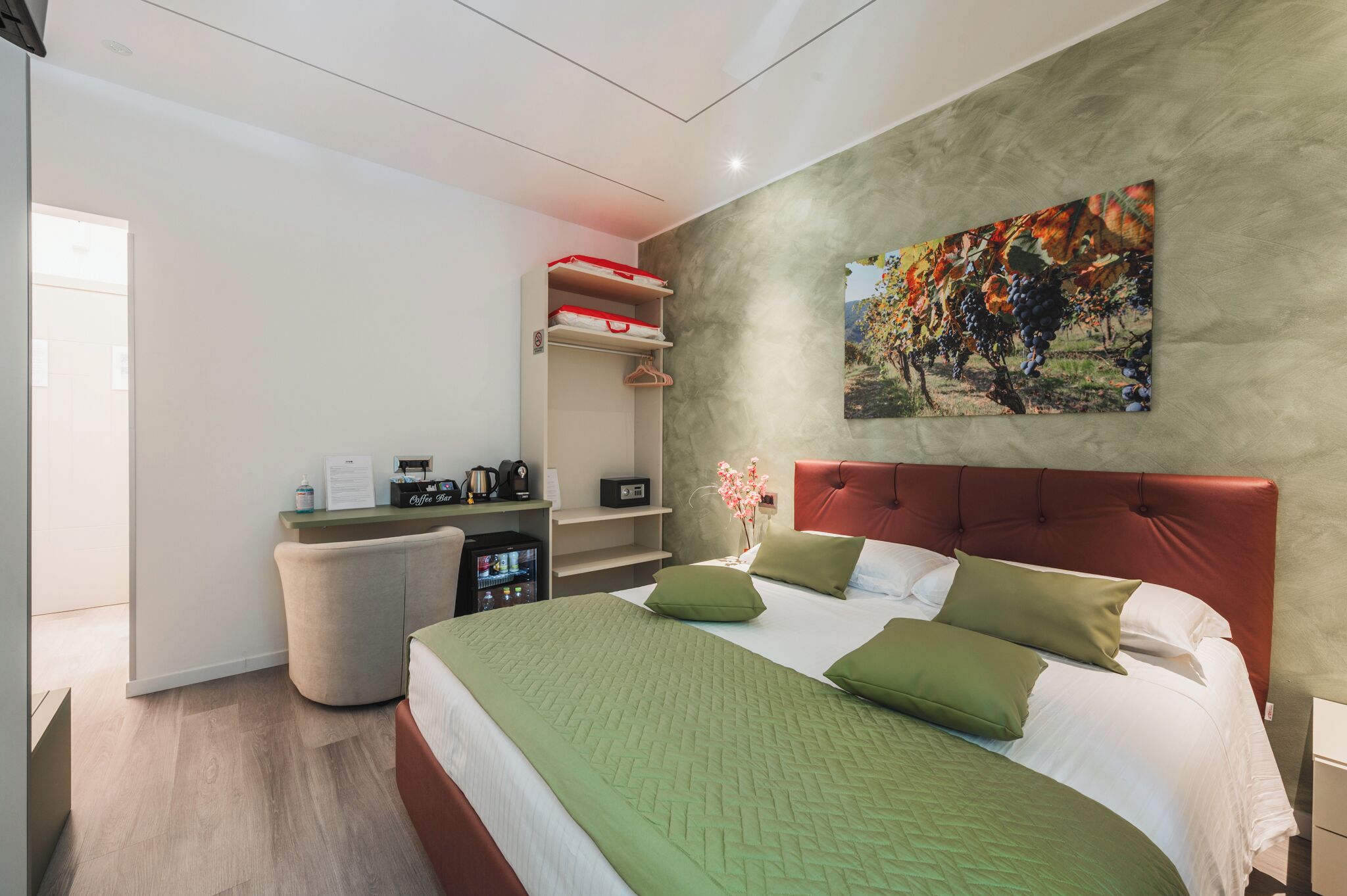 A modern hotel room with a large bed, green bedding, a red headboard, a wall-mounted painting, and a small kitchenette area.