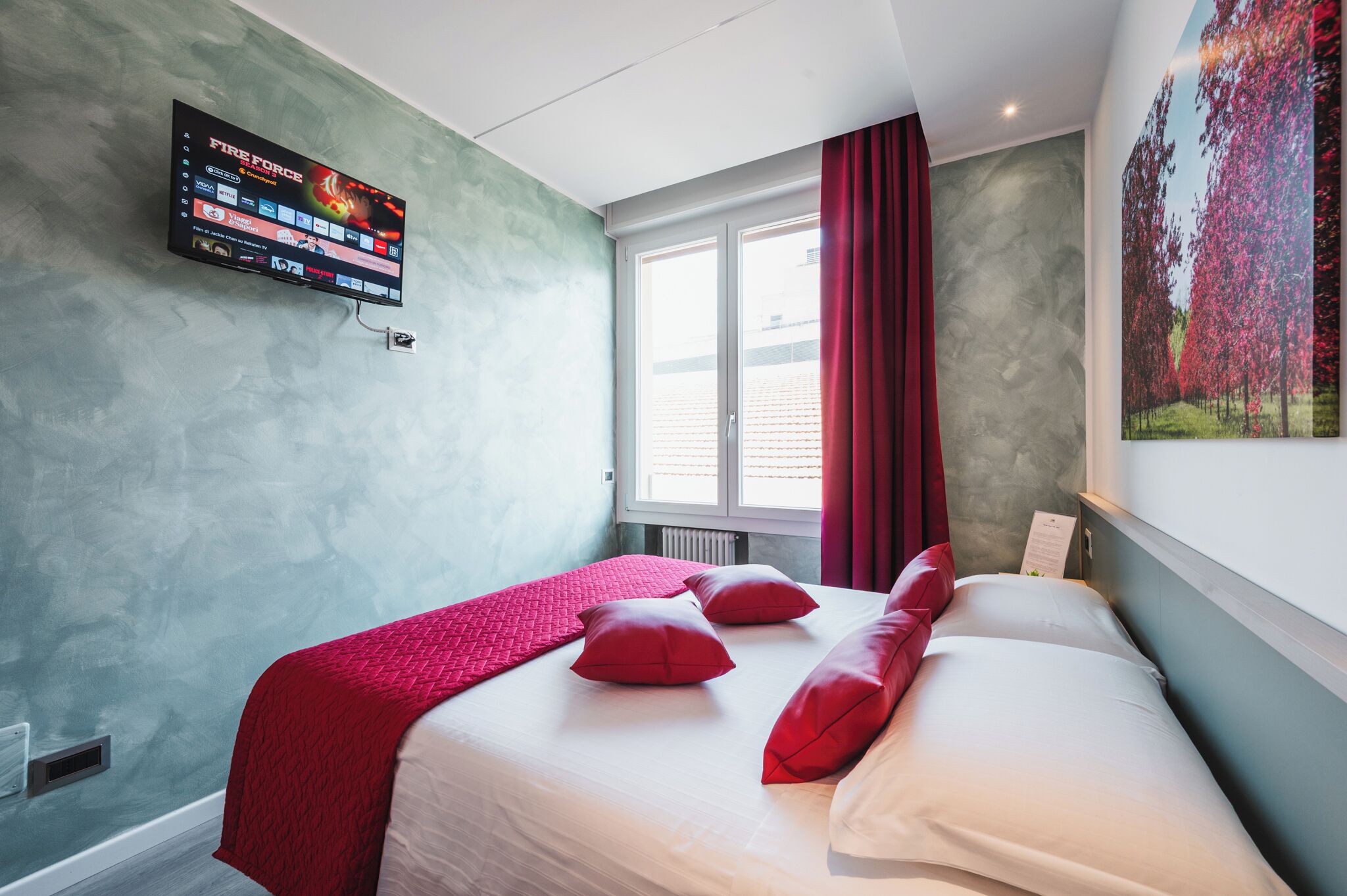 A hotel room with a bed, red pillows, a mounted TV displaying a game, a window with red curtains, and a wall-mounted painting.