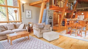 Un vaste espace de vie comprenant un canapé, un pouf et une table basse, une cheminée, des poutres en bois et un coin repas avec des chaises et une table.