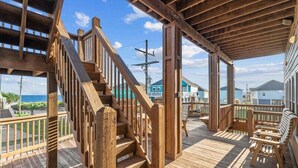 Een houten terras met een trap naar een overdekt gedeelte, buitenmeubilair en uitzicht op de oceaan en andere gebouwen.