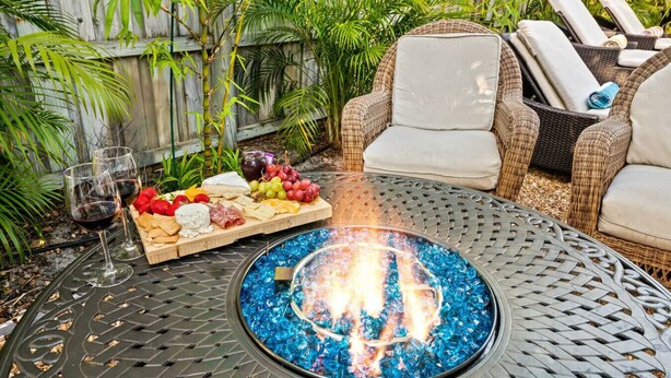 Een terras buiten met een ronde metalen tafel, rieten stoelen, een vuurkorf en een charcuterieplank.