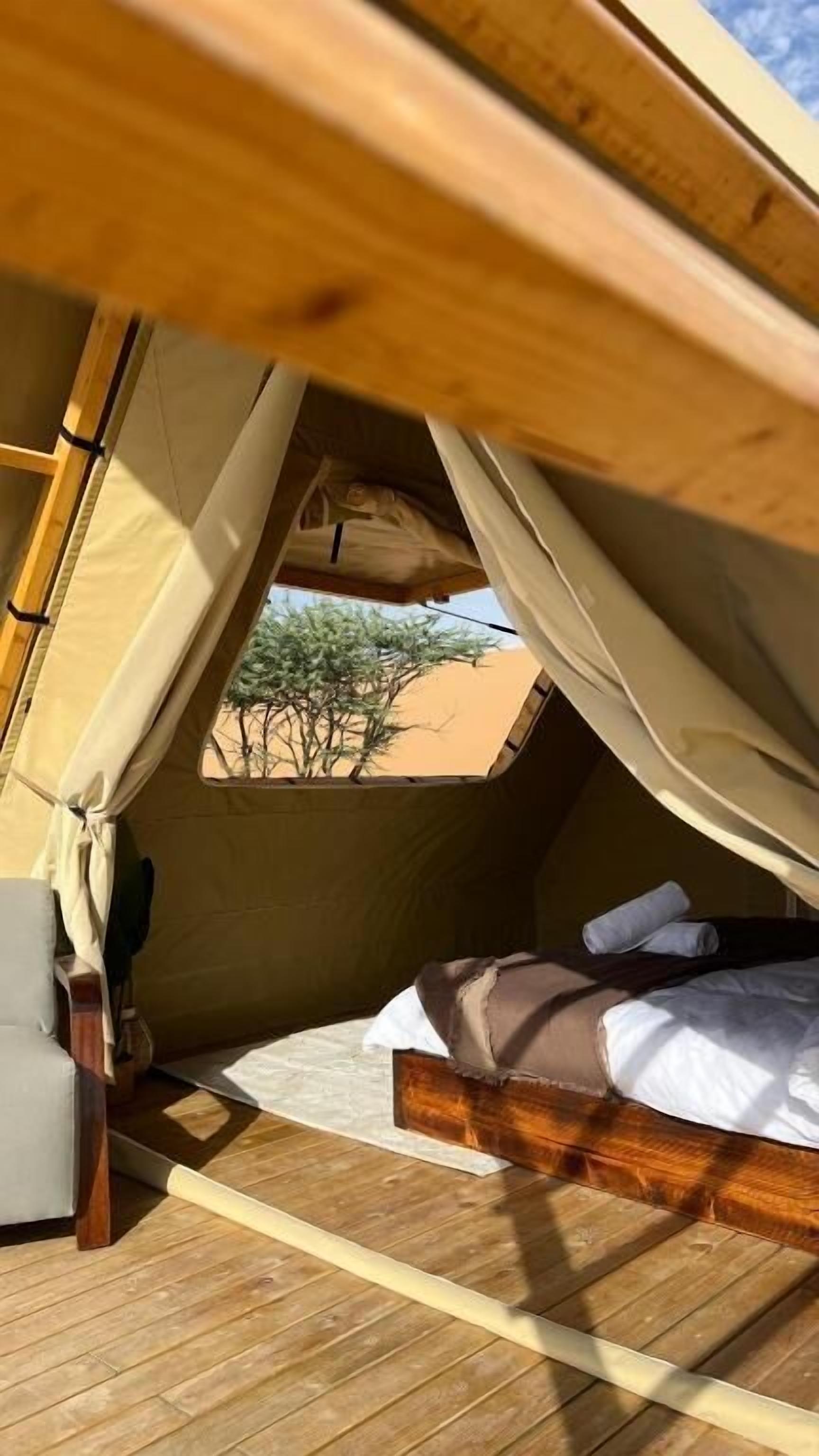 Une tente équipée d’un lit, d’une chaise et d’une fenêtre donnant sur des arbres et du sable.