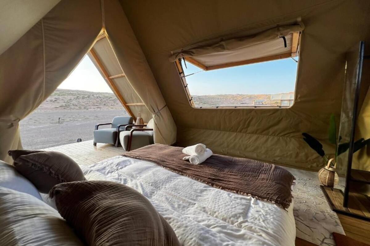 Una habitación con tienda de campaña, una cama, una pequeña mesa con sillas y una ventana con vistas a un paisaje desértico.
