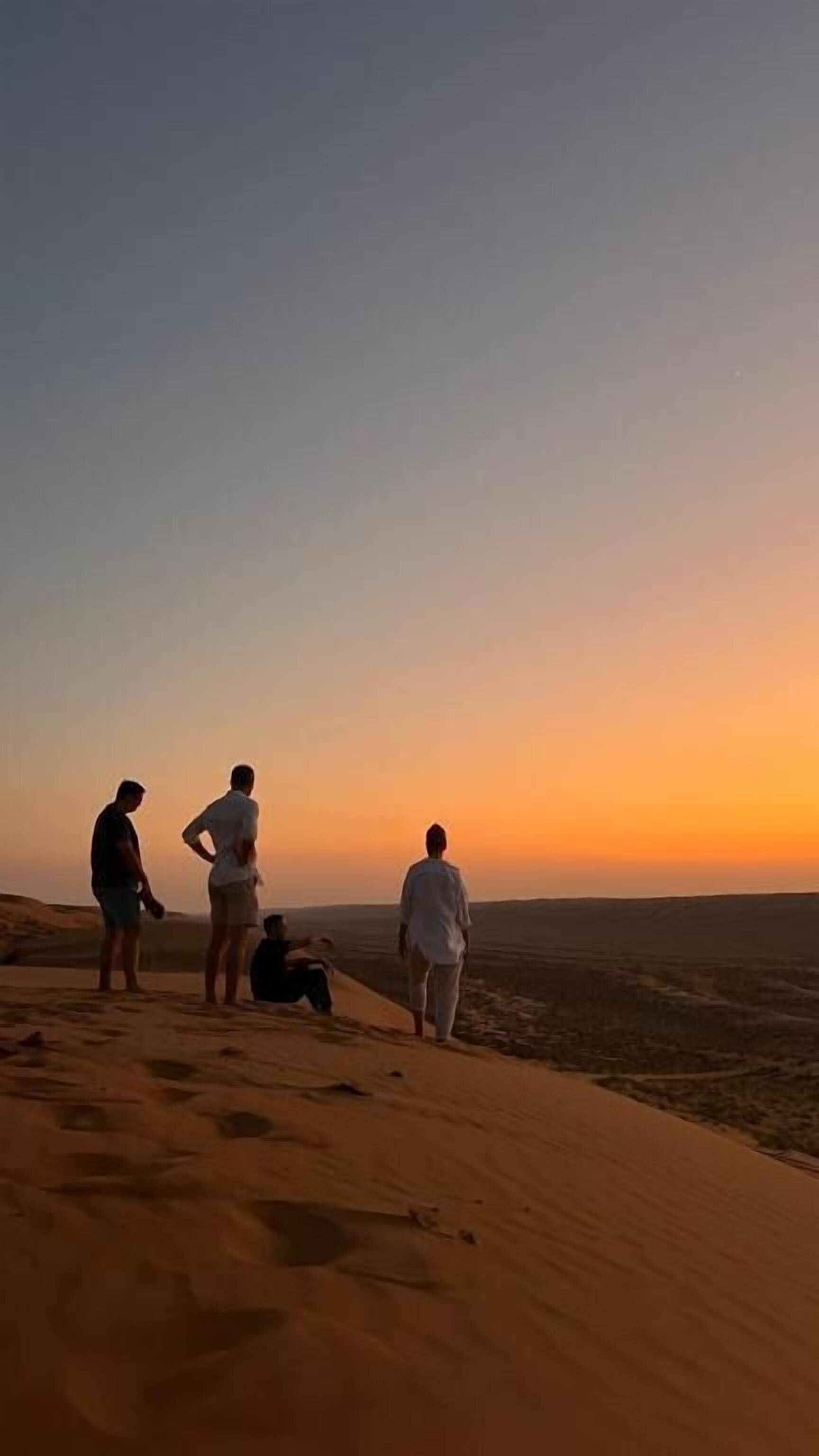 A group of people standing on a sand dune, overlooking a vast landscape during sunset.