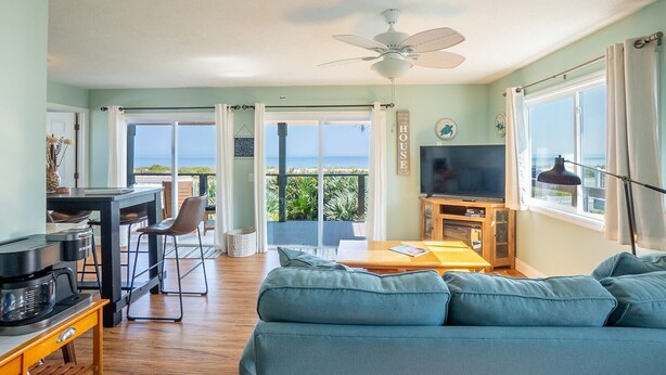 A living room with a sofa, a coffee table, a TV, and a ceiling fan. There is a balcony with a view of the ocean. - Flagler Beach Escape in the Sun (Flagler Beach)