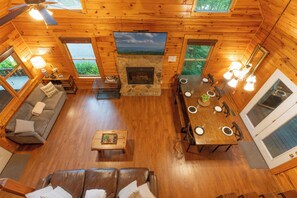A cabin interior with a stone fireplace, wooden walls, a sofa, a dining table, and large windows. - Epic Private Location*HotTub*FirePit*GameRoom (Pigeon Forge)