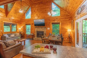 A cabin interior with a stone fireplace, wooden walls, a dining area, and a seating area with a couch and chairs. - Epic Private Location*HotTub*FirePit*GameRoom (Pigeon Forge)