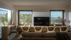 Uma sala de estar moderna com uma televisão de tela plana grande, um sofá e uma escrivaninha com cadeira. Há duas janelas com persianas e vista para edifícios e áreas verdes lá fora.