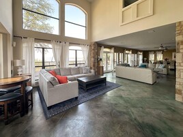 Amplia sala de estar con un sofá seccional grande, un sofá más pequeño y una mesa de centro. Hay un comedor con mesa y sillas. La habitación tiene ventanas grandes y vistas al exterior.