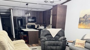 A living room with a beige recliner, a gray armchair, and a gray sofa. There is a kitchen in the background with a microwave, stove, and stools.