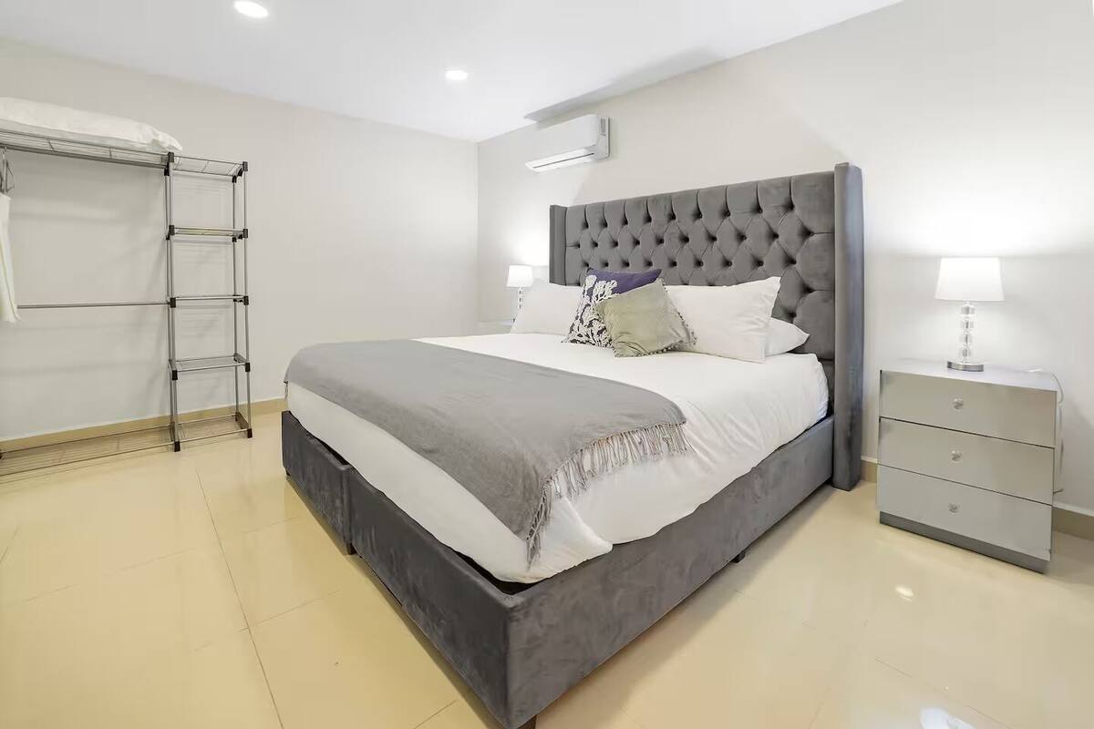 A modern bedroom with a grey tufted headboard, a grey bedspread, and a bedside table with a lamp.